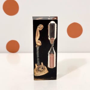 Peut inclure: Un bloc rectangulaire en résine noire contenant un téléphone à cadran miniature de couleur dorée avec un cordon en spirale et un sablier en verre transparent rempli de sable rose. Les objets sont incrustés dans la matière sombre et brillante.