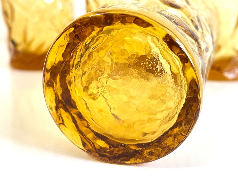 Op de afbeelding: Close-up van de bodem van een heldere amberkleurige glazen tumbler met een getextureerd oppervlak. Het glas heeft een dikke, afgeronde basis.