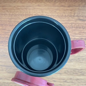 May include: A black and burgundy travel mug with a lid. The mug has a black interior and a burgundy lid and handle.