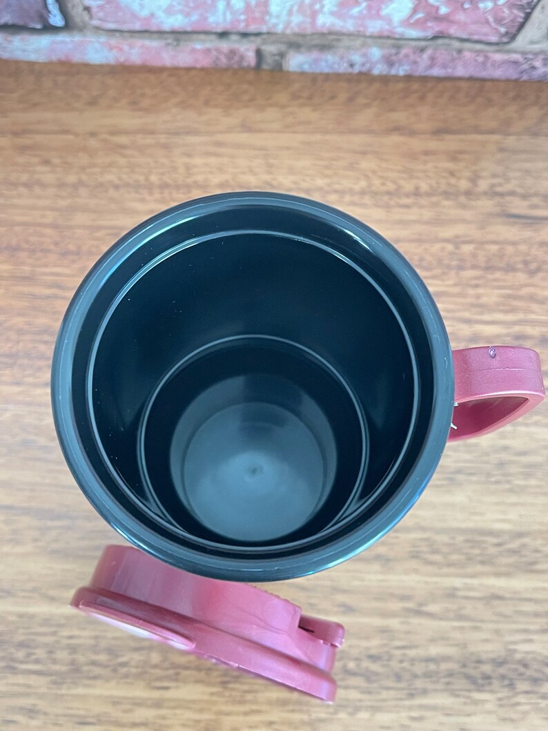 May include: A black and burgundy travel mug with a lid. The mug has a black interior and a burgundy lid and handle.