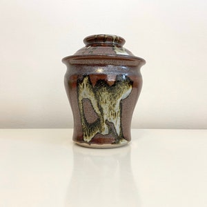 May include: A brown ceramic jar with a lid, featuring a mottled glaze with brown, white, and green accents. The jar has a rounded body and a slightly flared lip.