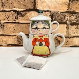 May include: A ceramic tea-for-one set featuring a speckled off-white glaze. The top cup depicts an elderly person with glasses, while the bottom teapot shows a person in an orange sweater and yellow apron knitting. Two tea bags sit in front.