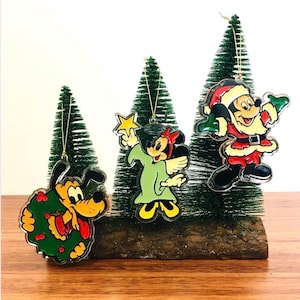 May include: Three Christmas tree decorations featuring Disney characters: Pluto with a wreath, Minnie Mouse in a green robe holding a star, and Mickey Mouse as Santa Claus. Each ornament hangs from a gold string.