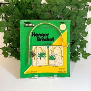 May include: A green and yellow package for a "Hanger Bracket" with the text "hold-all" at the top. The package shows illustrations of how to use the bracket for hanging plants on walls and windows. The bracket itself is gold-coloured.