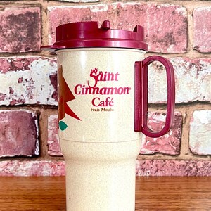 May include: A beige travel mug with a red lid and handle. The mug has a red and white logo that says "Saint Cinnamon Cafe" with the text "Frais Moulu" underneath.