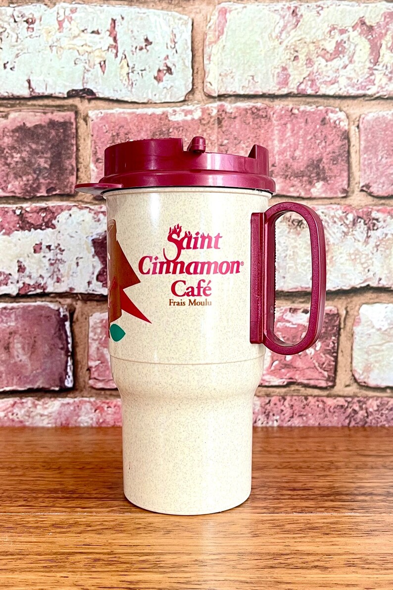 May include: A beige travel mug with a red lid and handle. The mug has a red and white logo that says "Saint Cinnamon Cafe" with the text "Frais Moulu" underneath.