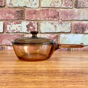 May include: A translucent amber-coloured glass saucepan with a matching glass lid. The cookware features a single long handle extending from the side and a circular knob on the centre of the lid, constructed from heat-resistant glass material.