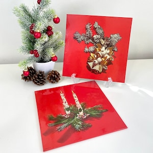May include: Two red square Christmas cards with a white background. The top card features a gold bow with white bells and a sprig of green pine. The bottom card features two white candles with a sprig of green pine.