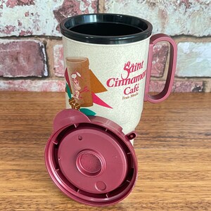 May include: A red and white travel mug with a black lid and handle. The mug has a graphic of a cartoon chef and the text "Saint Cinnamon Cafe Frais Mocha".