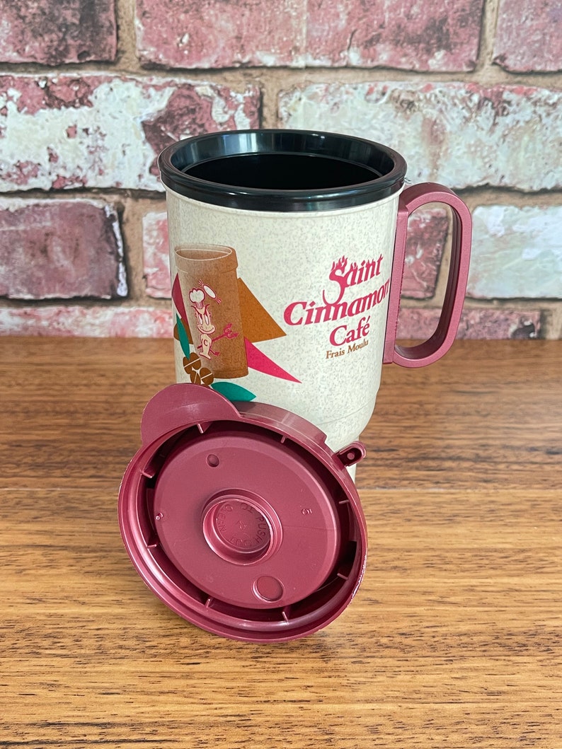 May include: A red and white travel mug with a black lid and handle. The mug has a graphic of a cartoon chef and the text "Saint Cinnamon Cafe Frais Mocha".