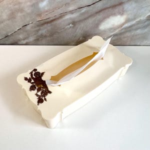 May include: Cream-coloured tissue box with a decorative cherub and floral design in dark brown. The rectangular box has a scalloped edge and a slot for tissue dispensing. A white tissue is partially pulled out.