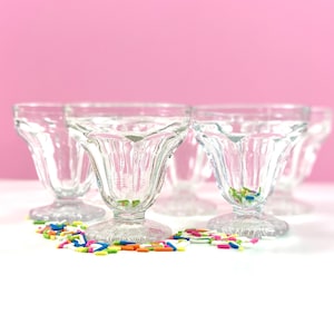 May include: A set of clear glass sundae dishes with a fluted design. The dishes are on a white surface with colorful sprinkles scattered around them. The background is a solid pink.