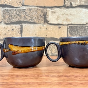 May include: Two black ceramic mugs with brown and yellow glaze. One mug has a brown and yellow stripe around the top, while the other has a brown and yellow stripe with a more abstract design.