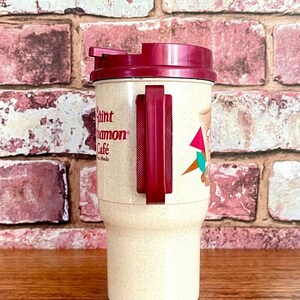 May include: A beige travel mug with a maroon lid and handle. The mug has the text "Saint Cinnamon Cafe" printed on the side.