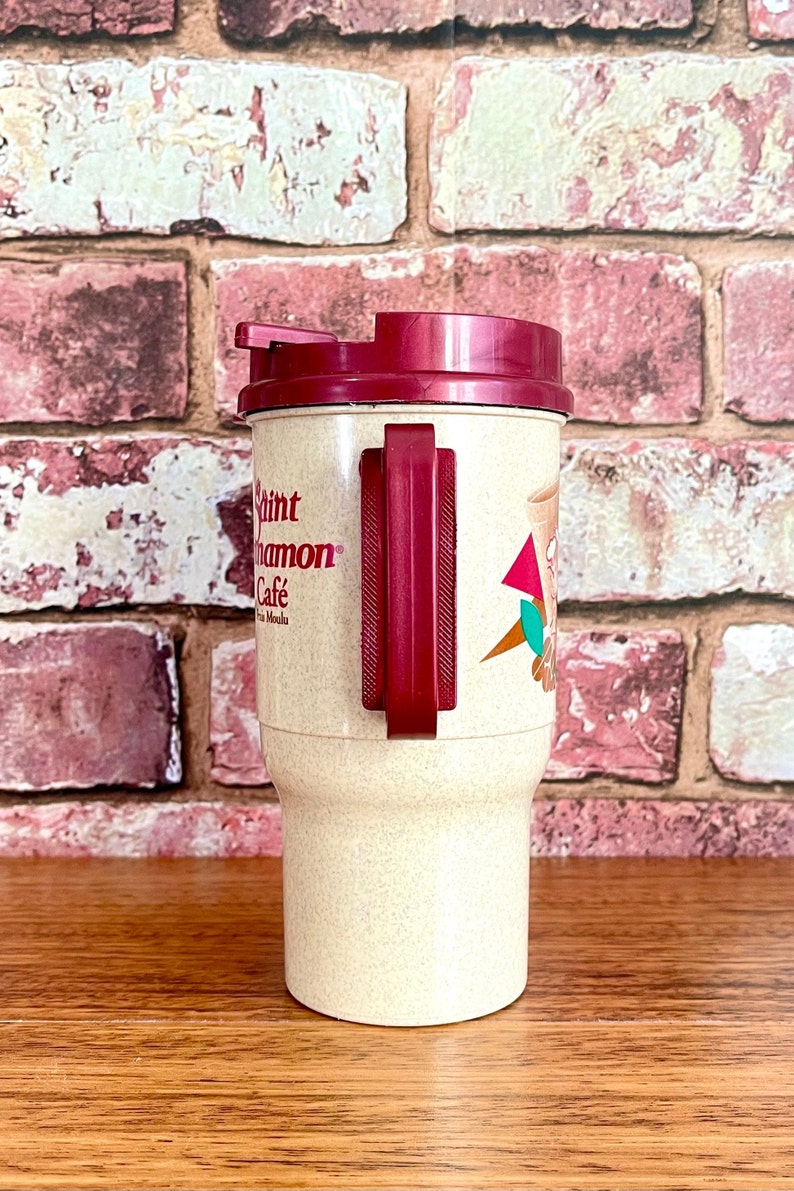 May include: A beige travel mug with a maroon lid and handle. The mug has the text "Saint Cinnamon Cafe" printed on the side.