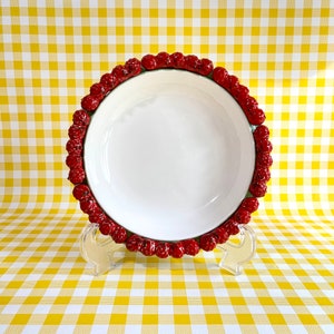 May include: A white ceramic bowl with a red berry design around the rim. The bowl is on a yellow and white checkered tablecloth.