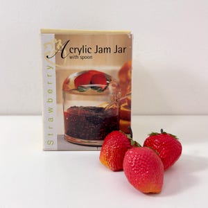 May include: A rectangular product box labeled Acrylic Jam Jar with spoon and Strawberry, featuring a clear jar with a strawberry-shaped lid filled with dark jam, accompanied by three realistic red strawberry replicas placed in front of the box.