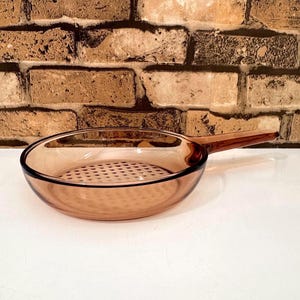 May include: An amber-coloured glass frying pan with a long handle. The pan has a round shape with a perforated base. The handle is attached to the side. The pan is on a white surface. The pan measures approximately 20 cm in diameter.