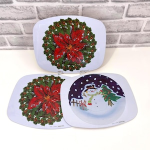 May include: Three white square plates with rounded corners. Two plates feature a central red poinsettia flower surrounded by a green holly wreath. The third plate displays a snowman wearing a hat and scarf next to a green Christmas tree.