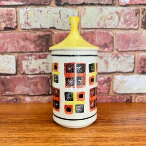May include: A white ceramic canister with a yellow lid and a geometric pattern in black, orange, and yellow. The pattern features squares and stars.