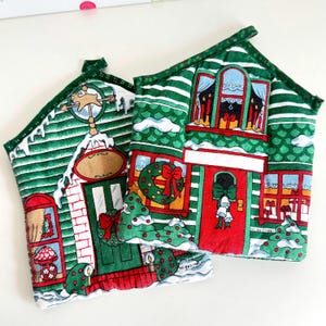 May include: Two green and white Christmas-themed pot holders shaped like houses with red doors and windows. The pot holders have a white background with green stripes and red accents. The text "No Bad Days" is visible on the bottom of the pot holders.