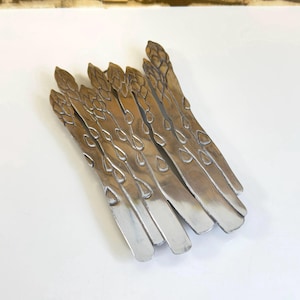 May include: A silver-toned metal asparagus sculpture with a textured surface. The sculpture is made of multiple pieces of metal that are joined together to form the shape of asparagus.