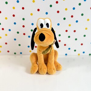 May include: A plush toy of Pluto, the Disney dog, sitting with a brown and green bow tie. He is orange with black ears and a black nose.