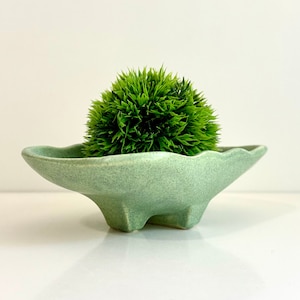 May include: A light green ceramic planter with a wavy edge and three legs. The planter is filled with a green artificial plant.