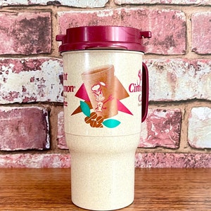 May include: A beige travel mug with a red lid and a cartoon character on the front. The mug has the text "Cinnamon Coffee" on the side.