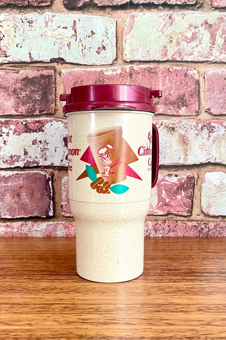 May include: A beige travel mug with a red lid and a cartoon character on the front. The mug has the text "Cinnamon Coffee" on the side.