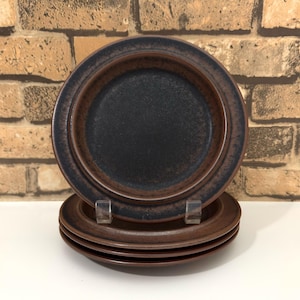 May include: A stack of four round ceramic plates featuring a dark brown centre that transitions into a deep black rim. The plates have a textured, matte finish with a raised edge, creating a simple, functional design for serving food.