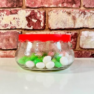May include: A clear glass jar with a red lid filled with green and white candies. The jar has a textured surface and is sitting on a brick background.