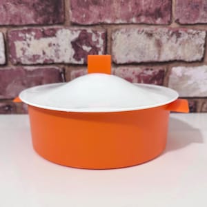 May include: An orange and white plastic food container with a lid. The container is round with a white lid and an orange handle. The container is sitting on a white surface. The background is a brick wall.