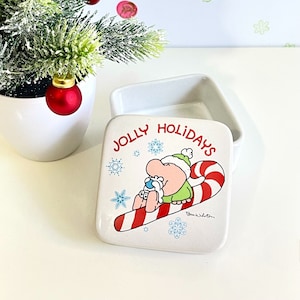 May include: White ceramic trinket box with a square lid. The lid features a cartoon character riding a candy cane with the words "Jolly Holidays" printed on the lid.
