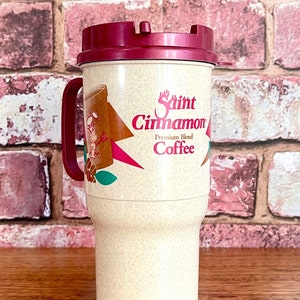 May include: A beige travel mug with a red lid and a Saint Cinnamon Coffee logo. The logo features a cartoon cinnamon stick with a face and a red star.