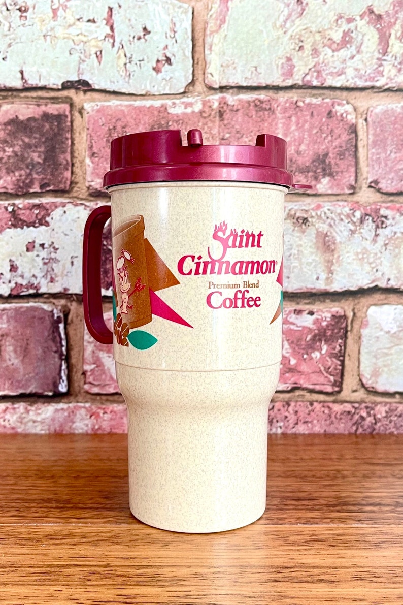 May include: A beige travel mug with a red lid and a Saint Cinnamon Coffee logo. The logo features a cartoon cinnamon stick with a face and a red star.