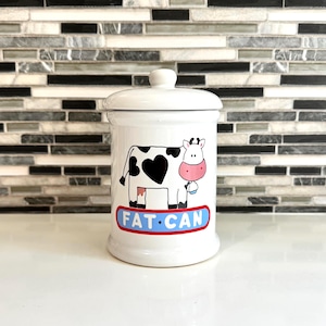 May include: White ceramic canister with a lid, featuring a cartoon cow design with a heart-shaped spot and the text "FAT CAN" in a blue and red oval. The canister has a rounded shape and a small knob on the lid.
