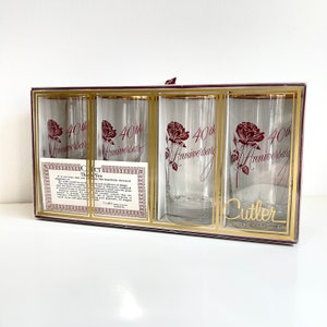 May include: Set of four clear glass tumblers with gold rims. Each glass features a red rose and the text "40th Anniversary". The glasses are packaged in a box with a gold and red label that reads "Cutler Signature Collection".