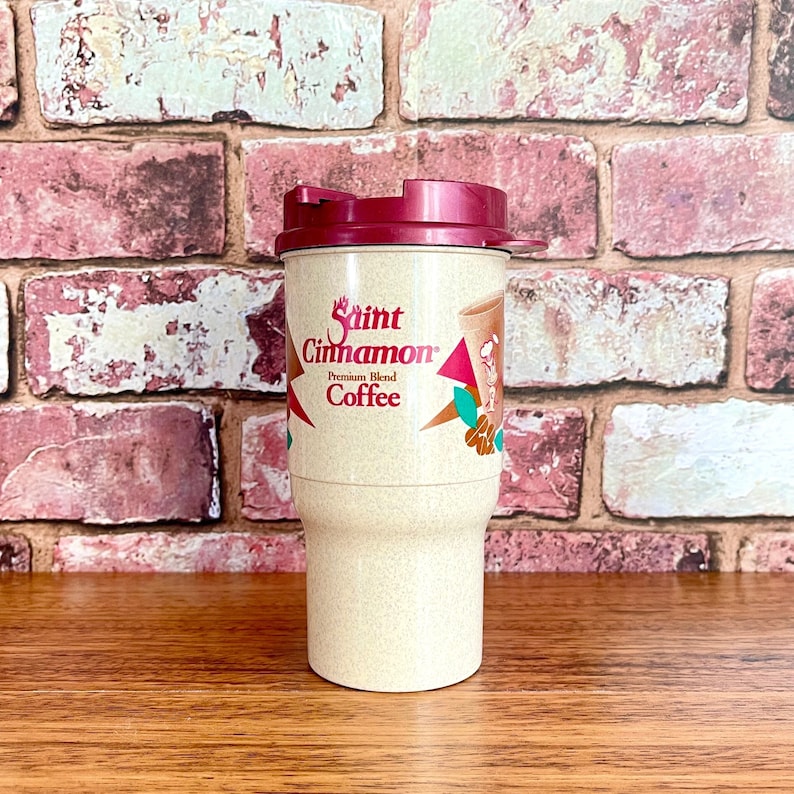 May include: A beige travel mug with a maroon lid and a Saint Cinnamon Coffee logo. The logo features a red star, a cinnamon stick, and a coffee cup.