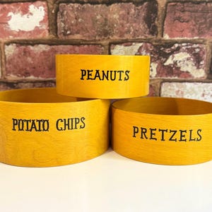Peut inclure: Trois bols à collation en bois jaune avec du texte noir. Les bols sont étiquetés "PEANUTS", "POTATO CHIPS" et "PRETZELS". Les bols sont empilés en forme de pyramide sur un fond de mur de briques.