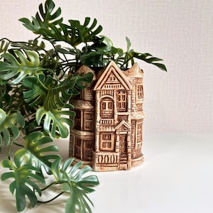 May include: A brown ceramic house-shaped plant pot filled with green leafy plants. The pot features detailed architectural elements such as windows, doors, and roof tiles. The plant's leaves are a vibrant green, providing a contrast to the pot's colour.