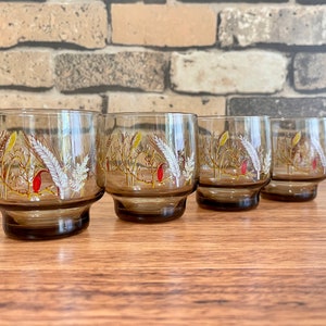 May include: Set of four brown glass tumblers with a white and yellow floral design. The glasses have a rounded bottom and a wide mouth.