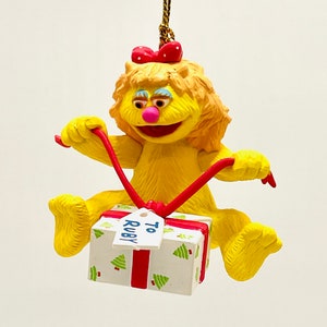 May include: A yellow character Christmas ornament with a red bow and a gift box. The gift box is white with green Christmas trees and a red ribbon. The tag on the gift box says "To Ruby".