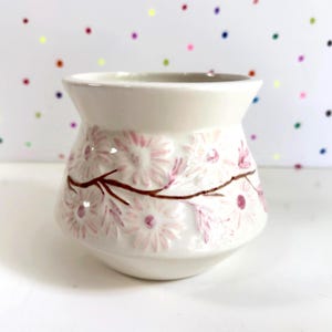 Peut inclure: Un vase en céramique blanc avec un dessus large et une base arrondie. Le vase est décoré de fleurs roses en forme de marguerites et de branches brunes. Le fond est blanc avec des pois colorés.