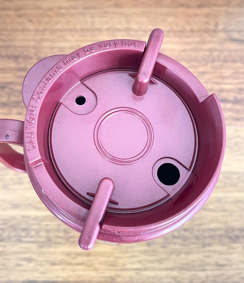 May include: A maroon travel mug with a lid that has a warning label that reads "CAUTION: Contents may be very hot."