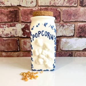 May include: White ceramic popcorn container with a blue and white design. The container has a cork lid and says "POPCORN" in blue lettering. A small pile of popcorn kernels is in front of the container.