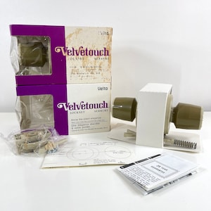 May include: Two boxes of a brown and white door lockset with the brand name "Velvetouch" printed on the front. The boxes are on a white surface. The lockset is a silent elegance door lockset. The lockset comes with a 25-year guarantee.