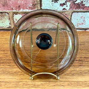 May include: A brown glass lid with a knob on top. The lid is sitting on a gold metal stand.