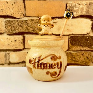 May include: A beige ceramic honey pot with a teddy bear lid and a wooden honey dipper with a bee design. The pot has the word "Honey" written on the front in brown script. The background is a brick wall.