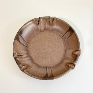 May include: A brown ceramic dish with a scalloped edge and a glazed finish. The dish has a light brown colour with darker brown streaks.